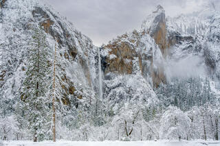 Winter photograph of Bridalveil Fall flowing through snow-covered cliffs with mist and pine trees in Yosemite National Park.