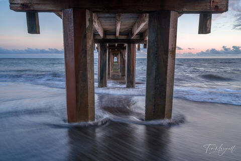 Fine-art photograph taken beneath Waimea Pier on Kauai’s west coast, showing waves flowing between pier pilings in a long exposure at dusk.