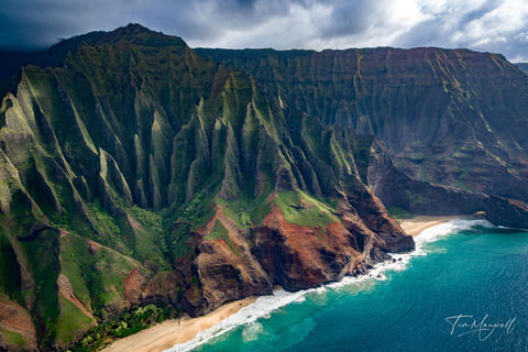 Fine-art aerial photograph of the Na Pali Coast on Kauai, showing dramatic green cliffs, turquoise ocean, and secluded beaches beneath moody skies.