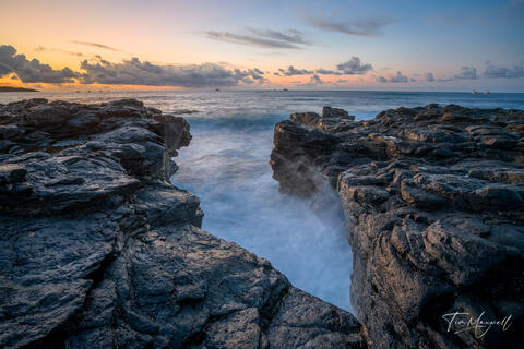 Fine-art photograph of sunrise on Kauai’s volcanic coast, showing waves rushing through a narrow lava rock channel framed by soft pastel skies.