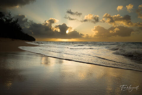 Fine-art photograph of sunrise at Gillen’s Beach, Kauai, with golden light streaming through clouds and reflecting on the wet sand.