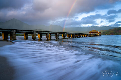 Fine-art photograph of a rainbow over Hanalei Bay on Kauai’s North Shore at sunrise, featuring the historic Hanalei Pier and soft ocean waves.