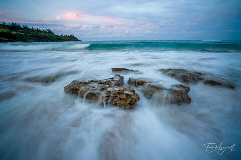 Fine-art photograph of waves flowing over coastal rocks at sunset on Kauai, with long exposure motion and pastel sky colors.