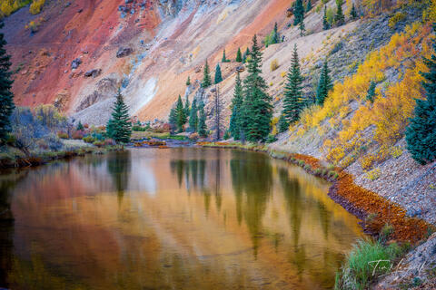 Fine art landscape photograph of colorful mineral slopes, evergreens, and autumn foliage reflected in still water along the Million Dollar Highway in Colorado.
