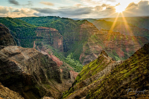 Fine-art photograph of sunrise over Waimea Canyon on Kauai, showing sun rays streaming across red cliffs and deep green valleys.